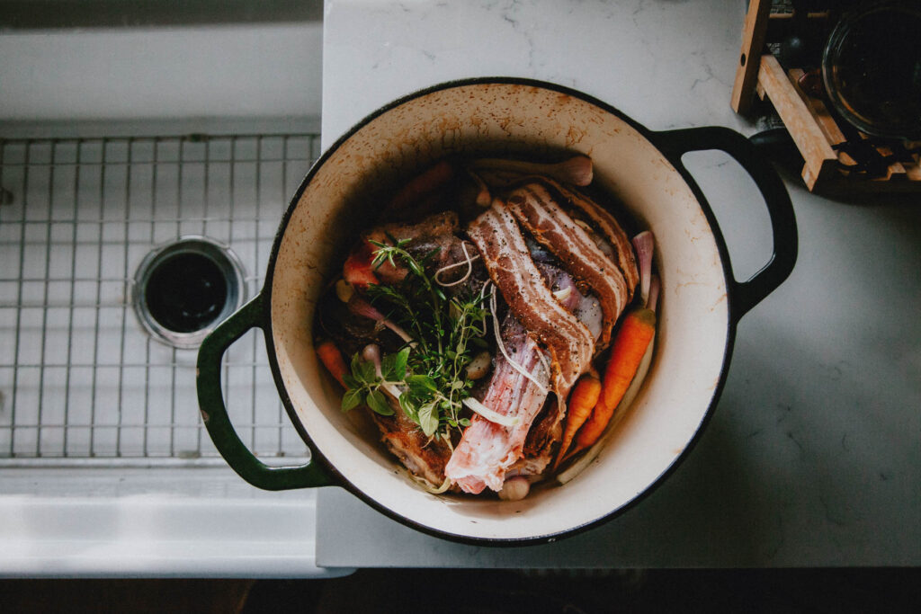 A dutch oven filled with goat meat wrapped in bacon rests on a marble countertop.