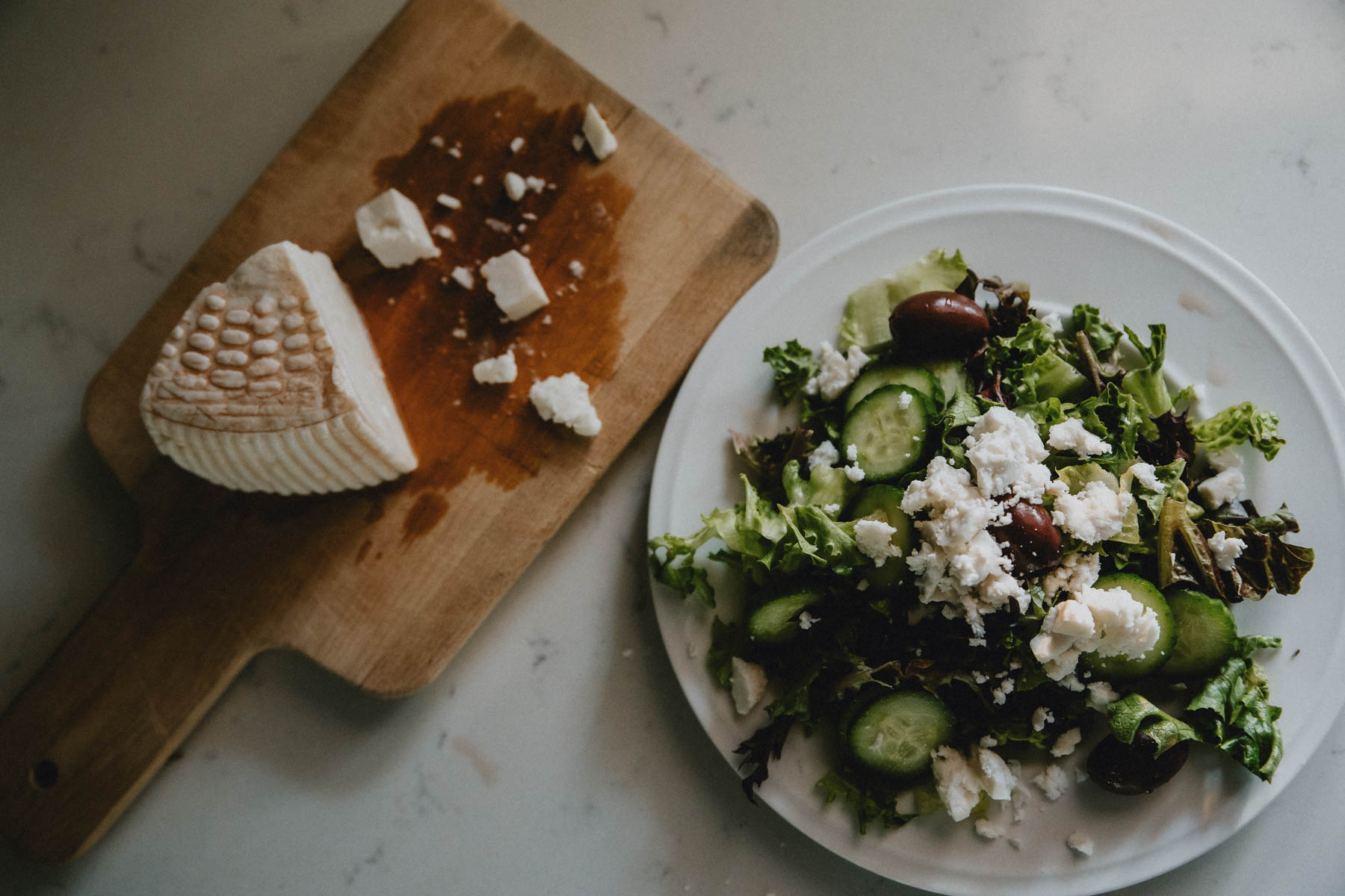A wedge of goat milk feta cheese sits next to a Greek salad topped with crumbled feta