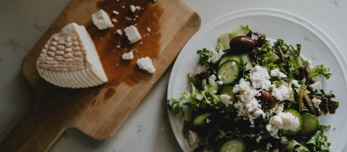 A wedge of goat milk feta cheese sits next to a Greek salad topped with crumbled feta